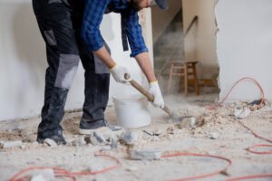 Close up shot of the legs of a man in a protective suit with a shovel in hand next to a bucket of debris left over from a house renovation.